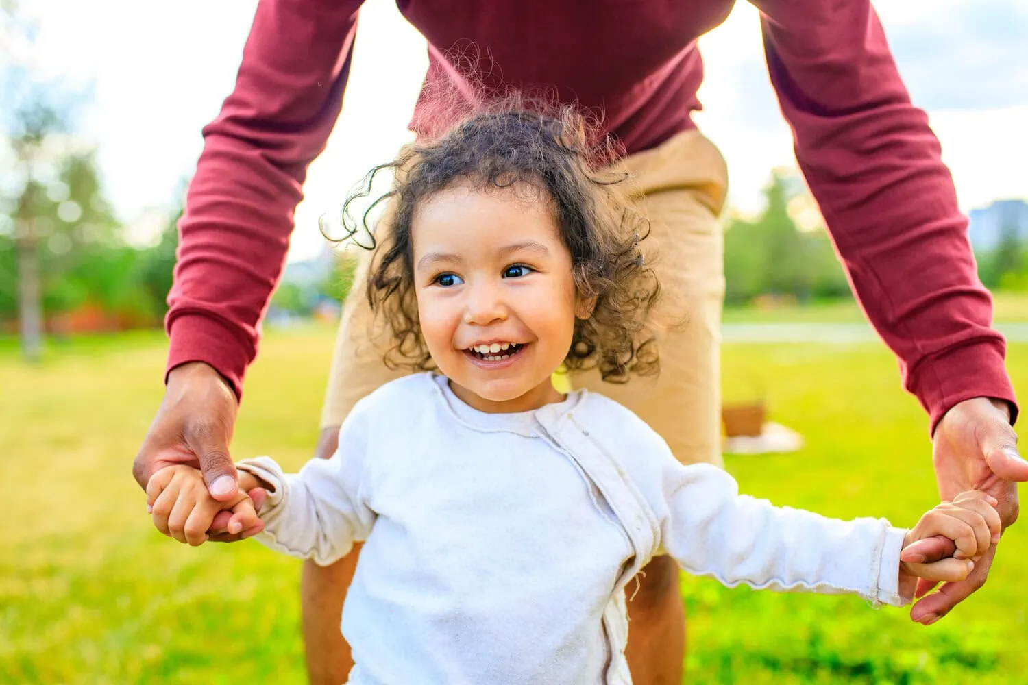 A child smiles while an adult holds both of their hands outdoors.