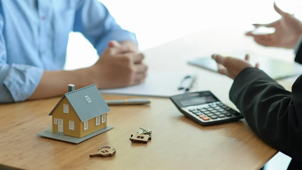Two people discuss insurance at a table with a model house and calculator.